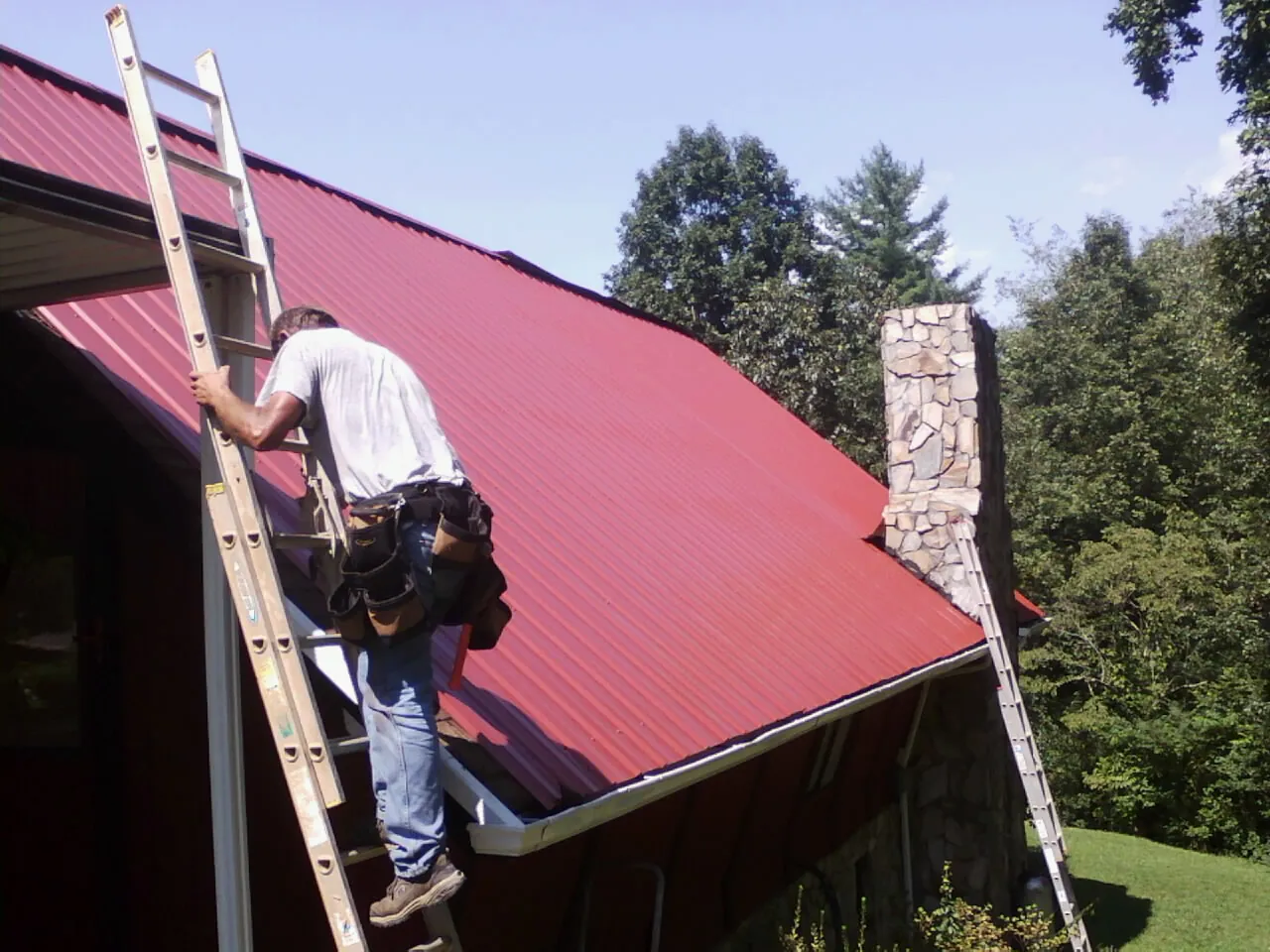 Roofer on a ladder beside a red metal roof and stone chimney — Soffit & Fascia Repair by Roofing Brothers in Carey